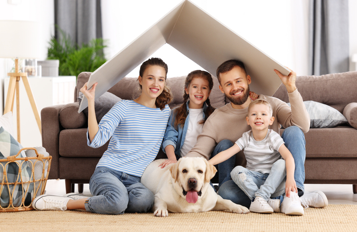 Young family with kids and dog holding roof over heads and smiling happily while sitting in living room of new apartment