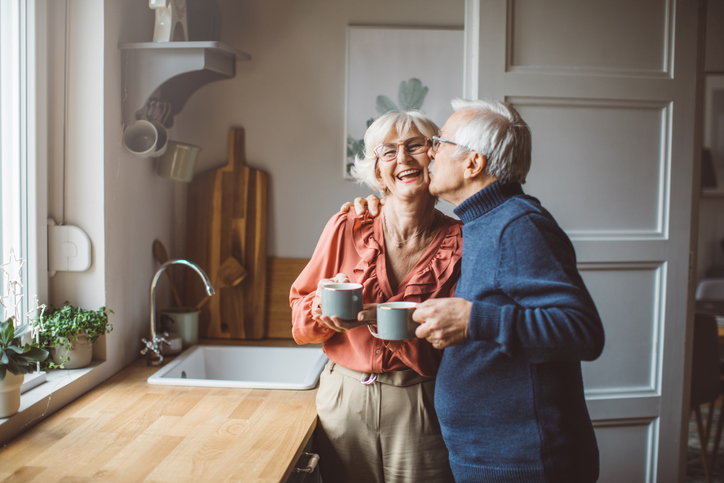 Senior couple at home standing in front of kitchen window and drinking tea or coffee.