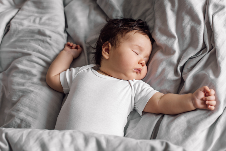 Portrait of baby in sleepwear lying under gray blanket on bed at home. Sleeping on back infant child in bedroom, see sweet dream. Grey background, free copy space. Childcare and healthy sleep concept